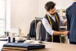 Tailor measuring a navy suit jacket in a bright shop with sewing tools and fine textiles on display.